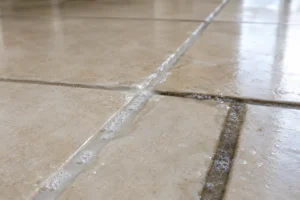 Pale cloudy cleaning solution pooled in the grout lines of a beige tile floor, with one treated line already lighter than the untreated line beside it