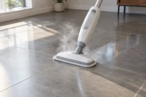 Upright steam mop releasing a plume of steam onto a gray porcelain tile floor, with a brighter damp strip visible behind it